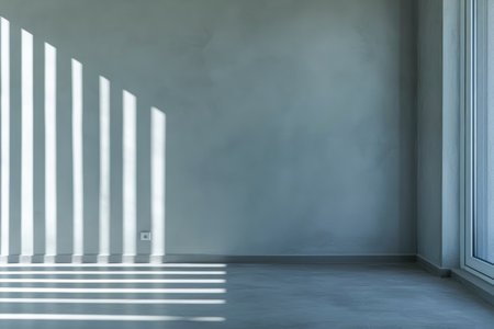 Bright sunlight filters through a large window, creating striped shadows on a bare, gray wall in a simple, spacious room. The floor is smooth and unadorned, enhancing the calm atmosphere.の素材