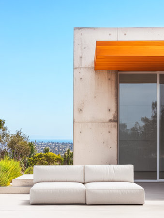 A stylish white sofa sits in a modern outdoor space, featuring a concrete wall and bright orange overhang, with a panoramic view of greenery and the distant city skyline.の素材
