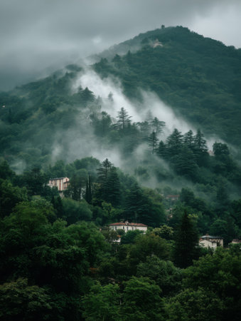 Mist blankets the mountains as clouds roll in, hovering over a serene village nestled among lush greenery in the early morning hours.の素材