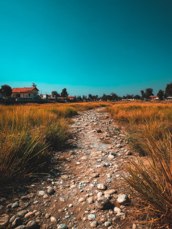 A winding dry riverbed surrounded by tall grass stretches towards a quiet village under a vibrant blue sky. Vegetation flourishes in warm sunlight, creating a peaceful scene in nature.の素材