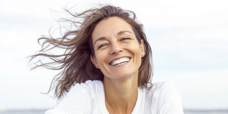 A joyful woman laughs joyfully while her hair dances in the wind by the water. The warm sunlight brightens her cheerful expression, adding to the serene outdoor atmosphere.の素材
