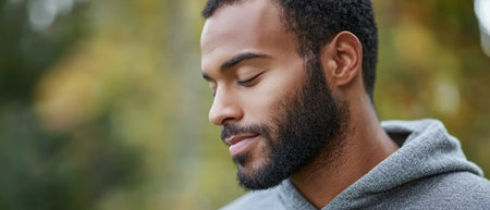 A man wearing a gray hoodie appears reflective while standing in a peaceful park setting. The surrounding trees and soft light create a serene atmosphere in the afternoon.の素材
