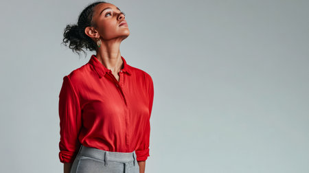 A woman stands tall wearing a bright red blouse and gray trousers, gazing upwards with a poised expression in a minimalist setting. Her confident stance highlights her elegance.の素材