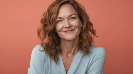 A woman with curly, red hair smiles warmly while wearing a light blue blouse. She is set against a soft coral background, conveying a sense of happiness and confidence.の素材