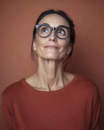 A woman with dark hair and glasses gazes upward thoughtfully against a warm, earthy orange wall. Her expression reflects curiosity and contemplation, showcasing a serene moment.の素材