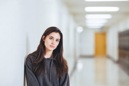 In a school hallway, a young woman in a hoodie leans against the wall, looking contemplative. Fluorescent lights illuminate the corridor, creating a calm atmosphere.の素材