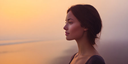 A young woman stands at the beach, gazing into the distance. The sun sets, casting warm colors across the sky and water. She appears deep in thought, embracing the peaceful surroundings.の素材