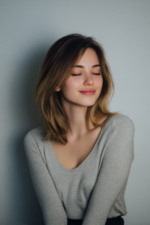 A young woman smiles gently with her eyes closed, exuding warmth and happiness. She wears a light gray sweater and poses against a minimal gray backdrop, creating a serene atmosphere.の素材
