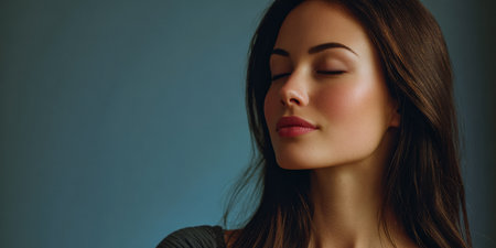 A young woman enjoys a peaceful moment indoors with her eyes closed. Soft lighting and a neutral background create a serene atmosphere, emphasizing her calm expression.の素材