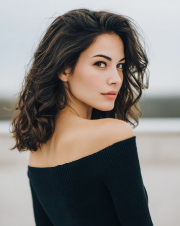 A woman stands outdoors, looking back at the camera with an alluring expression. Her dark curly hair frames her face, and she wears an elegant off-shoulder black top against a cloudy backdrop.の素材
