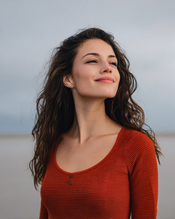 A young woman with wavy hair stands by the shore, smiling gently. She wears a red striped top and enjoys the fresh air on a cloudy day while looking content and peaceful.の素材