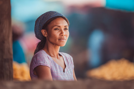 A woman wearing a traditional outfit sits quietly at a market stall. Surrounding her are blurred colors of fruits and vegetables, creating a lively atmosphere in the daylight.の素材