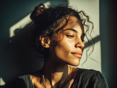 A woman with curly hair relaxes against a wall, illuminated by warm sunlight. Soft shadows create a peaceful atmosphere, enhancing her serene expression.の素材