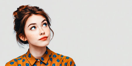 A young woman with brown hair tied up looks up thoughtfully. She wears a floral button-up shirt with a warm tone. The backdrop is simple and neutral, drawing focus to her expression.の素材