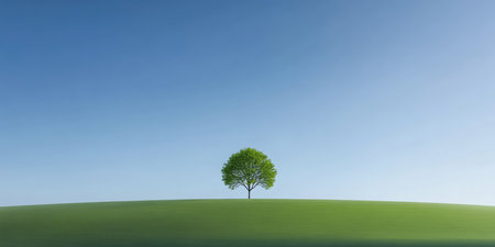 A vibrant green tree stands alone on a smooth grassy hill, set against a bright blue sky. This serene landscape captures the essence of nature's beauty in daylight.の素材