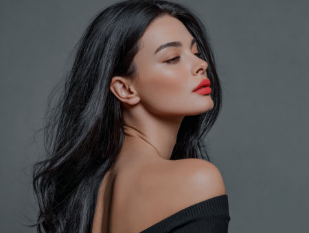 A woman with long dark hair poses gracefully, turning her head slightly to the side. Her lips are vibrant red, while she wears a chic black off-shoulder outfit. The background is simple and muted.の素材