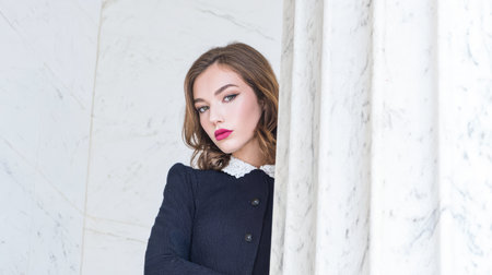 A model stands near a white marble wall. She wears a black outfit and has light makeup. The setting is bright with natural light coming in. The model looks directly at the camera.の素材