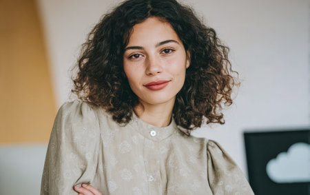 A woman stands in an indoor space with her arms crossed. She has curly hair and wears a light-colored shirt. The setting has simple decor and natural light.の素材