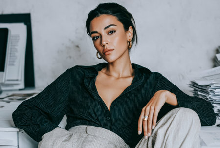 A woman with dark hair is sitting at a desk in an office. She wears a black shirt and light-colored pants. Papers and a computer are visible on the desk around her.の素材