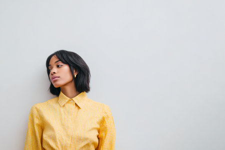 A woman stands against a light wall. She has shoulder-length dark hair and wears a yellow shirt. The setting is bright and simple, showing focus on her expression.の素材