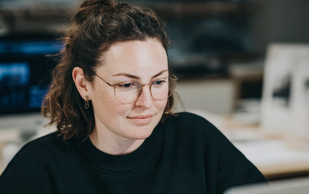 A woman sits at a desk in an office, looking at a computer screen. She appears concentrated on her work. Soft lighting fills the room in the late afternoon.の素材