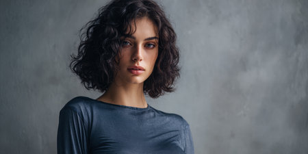 A woman stands in front of a gray wall with her curly hair styled naturally. She is wearing a fitted top and looks directly at the camera during a photo session inside a studio.の素材