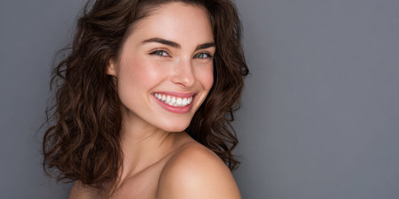 A woman with curly hair poses smiling while looking directly at the camera. She stands in front of a gray background with a natural expression on her face.の素材