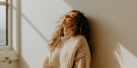 A woman stands against a wall in a well-lit room. Sunlight pours in from a nearby window. She has wavy hair and smiles softly, showing a moment of relaxation and comfort.の素材
