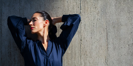A woman stands with her back against a concrete wall, her hands raised in her hair. She wears a dark shirt and has a confident expression under bright sunlight.の素材