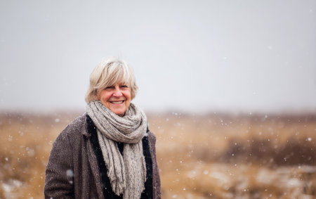 A woman stands in a snowy field smiling while snow falls around her. The scene shows tall grass in the background on a cold winter day.の素材