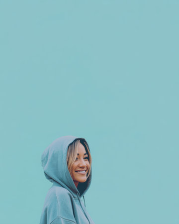 A woman stands with a slight smile, wearing a hoodie. She is near a light blue wall. The scene is bright and captures a casual moment in the day.の素材