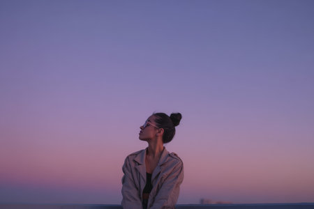 A person is sitting on a low wall looking up at the sky. The sun is setting, and the colors change from blue to purple and pink in a city background.の素材