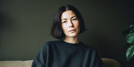 A woman sits on a couch with a serious expression. She looks directly at the camera. The setting is a room with simple decor and indoor plants.の素材