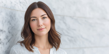 A young woman with brown hair stands by a marble wall. She faces the camera and maintains a direct gaze. The background is light and simple. Natural light brightens the scene.の素材