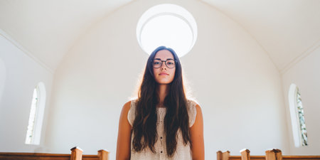 A young woman stands inside a well-lit room with a large round window above her. She looks directly at the camera with a serious expression. The light highlights her features.の素材