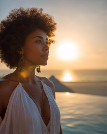 A woman with curly hair looks out over the ocean at sunset while standing next to a pool. The sun sets on the horizon and creates reflections on the water.の素材