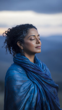 A woman stands outdoors on a mountain with her eyes closed. She is wearing a blue scarf and appears to be enjoying the fresh air during sunset. The sky has soft colors.の素材