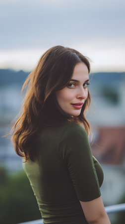 A woman stands outdoors on a balcony. She turns her head to look at the camera. Buildings are visible in the background. The sky is cloudy with a hint of sunset light.の素材