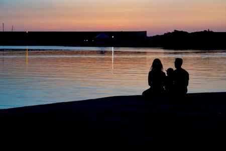 Silhouette of a man, woman, and child sitting by the water at sunsetの写真素材