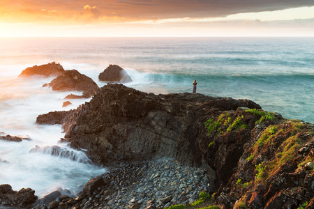 A woman is dwarfed in a majestic coastal scene as the sun rises and highlights the vibrant colours...