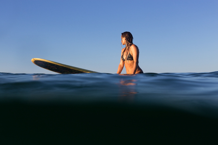 A beautiful female surfer sits on her surfboard in the evening light on the Pacific Ocean.の写真素材