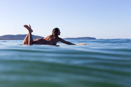 A beautiful female surfer is silhouetted by the evening sunlight as she lays on her surfboard in a...