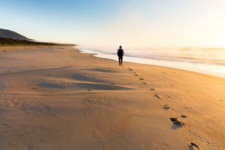 A person walking on a beautiful beach leaves a trail of footprints as the sunrise illuminates mist...