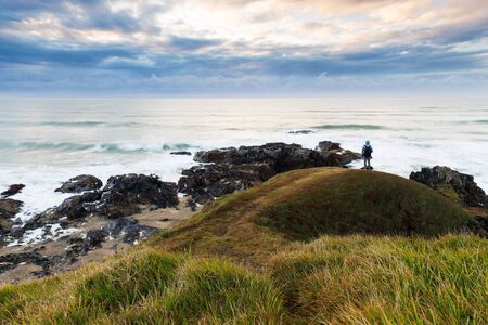 A person stands on a grassy headland and watches a storm pass on the horizon over a rough ocean.