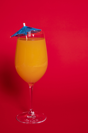 Orange drink with a blue umbrella in a wine glass set against a solid red background.の写真素材