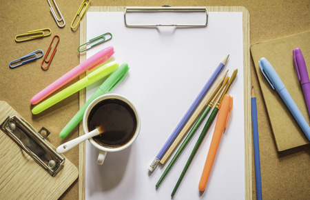 Stationery: clipboard, clips, pencils, color pens, paintbrush, notebook and coffee on wooden table topの写真素材