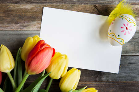 Colorful red and yellow tulips and Easter egg, with a blank card on rustic wooden table.の写真素材
