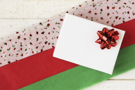 Small white gift box with red bow on colorful red and green tissue paper.