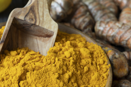 Close up of turmeric powder in bowl with scoop and turmeric roots in background.