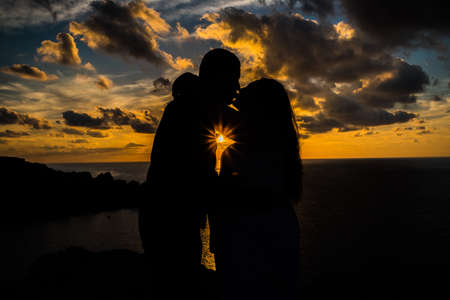 Silhouette of a man and a pregnant woman. A loving couple kissing each other during sunset with clouds and sea at the background. Horizontal. Side Viewの写真素材